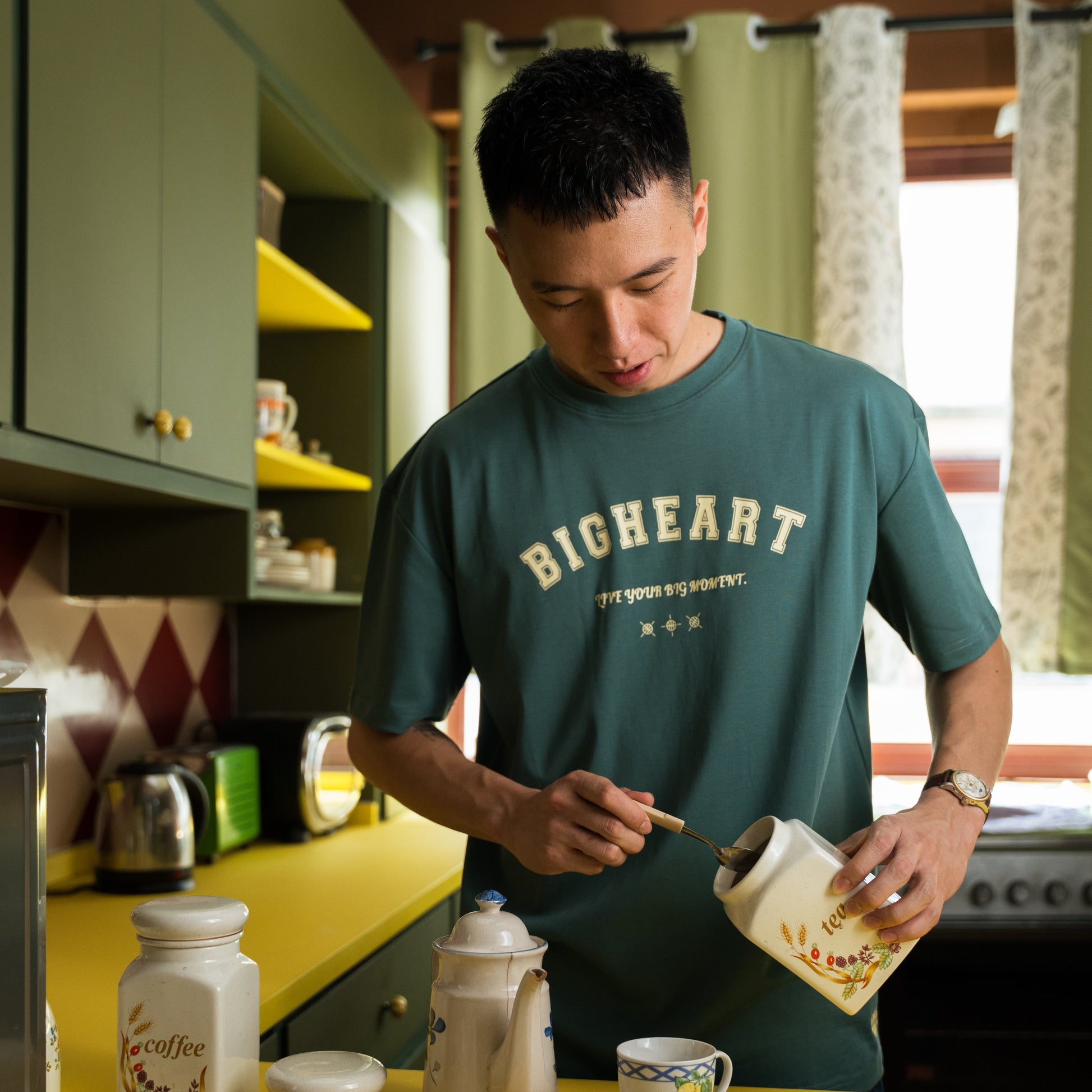 Man in a kitchen preparing tea, wearing a green 'BIGHEART' t-shirt.